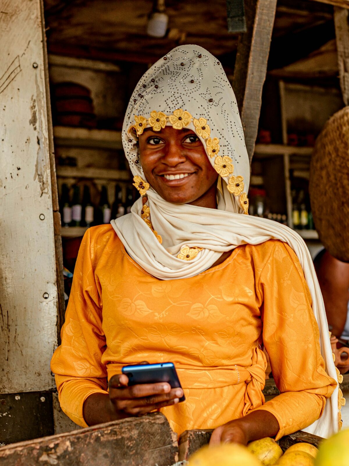 A smiling woman in an orange dress and a cream headscarf stands at a wooden market stall holding a mobile phone, with fruit on the counter in front of her.